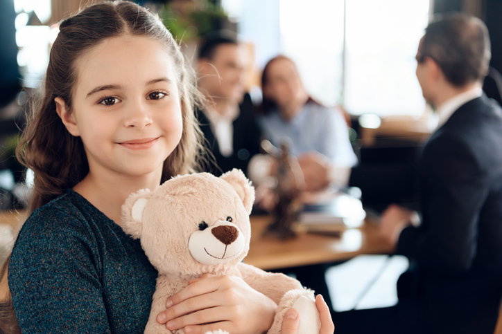 A young girl stands with a teddy bear and smiling at camera. Beautiful girl with parents stands in the office of a lawyer.